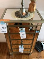 Front view of the mirrored jewelry armoire showing three drawers with metal pull knobs and the mirrored top surface with items on top.