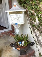 Front view of white mailbox showing embossed horse rider and 'LETTERS' sign along with wrought iron planter containing fern and small ornaments.
