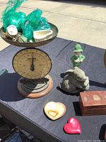 Photo showing Salter scale with brass tray holding various bubble-wrapped china pieces, ceramic figurine of boy in green, metal dog figurine, wooden box, and two decorative stone hearts on a black cloth