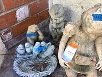 Photo showing six cement statues grouped in a corner against a brick and stucco wall. Includes a baby statue, a girl statue, a small dog statue, and three birds on a nest-shaped base with leaves inside.