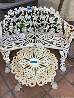 White wrought iron loveseat and round table with ornate grapevine and leaf designs, featuring claw feet legs, placed outdoors on tile flooring.