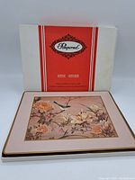 Photo of four acrylic placemats featuring floral and bird design on a pink background with cream border, arranged on a white surface with original box behind.