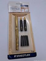 Original sealed packaging showing the gold tone pen, two black nib holders with gold nibs, and four ink cartridges labeled Staedtler Calligraphy Set.