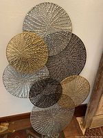 Overall view of the metal wall decor showing seven overlapping circular plates in silver, gold, and bronze finishes with a perforated pattern.