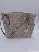 Front view of Michael Kors monogram tote handbag showing beige and brown MK pattern, two tan leather shoulder straps, and gold-tone MK logo charm.