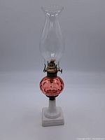 Full view of the oil lamp showing the white porcelain base, pink glass reservoir, brass wick holder, and clear chimney.