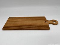 Full view of rectangular wooden cutting board with handle and hole, showing natural wood grain and branded mark on handle.