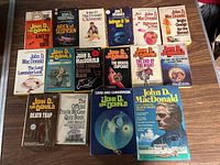 Top view of 14 John D. MacDonald paperback books arranged in rows, showing various cover titles and artwork.