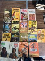 Top view of seventeen vintage Western theme paperback novels arranged laid flat on a wooden surface. Covers show colorful classic Western illustrations with cowboys, horses, and desert scenes.