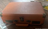 Closed brown faux leather briefcase style portable record player with metal corner protectors and handle.