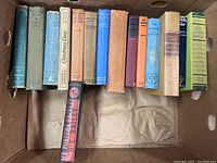 Photo of vintage books in a box, mostly hardcover with cloth bindings, titles visible include Trader Horn, Christmas Days, Out of Africa, The Rich, and others.