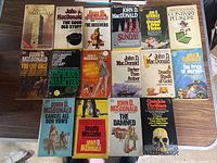 Photo showing 16 John D. MacDonald paperback novels arranged on a wood surface, titles clearly readable.