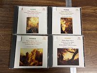 Four CD jewel cases arranged on table, featuring Weber Piano Music Vol. 1-4 with Alexander Paley piano performance. Each cover has classical paintings and text.