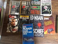 Photo showing nine paperback books by Ed McBain with various titles and cover art laid on a wooden surface.