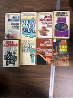 Seven vintage paperback John D. MacDonald books laid out on a wooden floor showing front covers: 'Dress Her in Indigo,' 'Nothing Can Go Wrong,' 'The Scarlet Ruse,' 'No Deadly Drug,' 'Murder for the Bride,' 'Cinnamon Skin,' 'The Neon Jungle'. Covers show some wear and creasing.