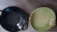 Top view of black clay bowl with two round handles and glossy finish beside green textured bowl with sculpted handles.