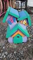 Front view of the wooden birdhouse showing the three joined house sections with green roofs, colorful windows and door on a ground bed of wood chips.