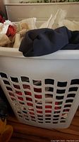 Front view showing white plastic laundry hamper filled with an assortment of vintage clothing and linen pieces including dark fabric on top and white lace underneath