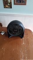 Black Honeywell electric fan sitting on a wooden table, showing full front and part of power cord.
