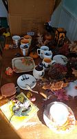 Overview photo of assorted vintage Halloween mugs, plates, and fall decor spread on a table with a cardboard box in background.