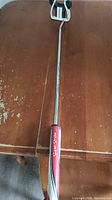 Full length of the putter showing the metallic shaft and red Super Stroke Slim 3.0 grip on a wooden surface.