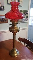 Full view of tall Victorian brass oil lamp on wooden table with red glass chimney.