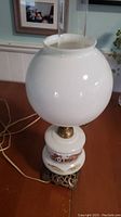 Front view of the lamp showing the white milk glass globe shade and middle section with scenic painting, on a wooden surface.