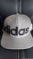 Front view of grey Adidas snapback hat with large black embroidered Adidas logo placed across front.
