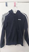 Front view of black Adidas zip-up hoodie showing white three-stripe sleeve detail, front zipper, and Adidas logo on chest.
