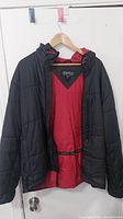 Front view of black Eddie Bauer men's quilted hooded jacket with red lining, hanging on wooden hanger against white door.