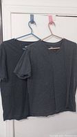 Photo of two dark gray V-neck T-shirts hanging on hangers, showing the front sides, one on a blue hanger and one on a pink hanger.