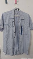 Front view of blue and white thin striped men's button-up shirt on hanger showing collar, buttons, pockets and short sleeves.