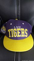 Front view of LSU Tigers purple and yellow snapback hat with large embroidered text and mascot logo on the side.