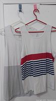 Two men's muscle shirts on hangers hanging on a white wall. One shirt is white with a gray bottom and chest pocket, the other has red and blue stripes.