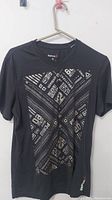 Front view of black Reebok t-shirt showing graphic design across chest with abstract patterns and text.