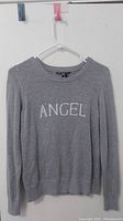 Grey knitted sweater with 'ANGEL' text on hanger against white background