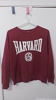 Front view of maroon women's sweatshirt with Harvard text and crest emblem on front, hung on a white hanger.