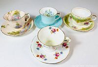 Top view showing four Aynsley bone china tea cups and saucers with distinct floral and color patterns including pink scenic, blue embossed, green floral Victorian and white floral sets.