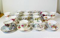 Front view of 16 tea cups and saucers arranged in four rows, showing diverse floral patterns and trimmed edges.
