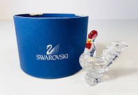 Clear Swarovski crystal rooster figurine with red and yellow accents on comb, wattle, and beak shown beside the original blue box with Swarovski swan logo.