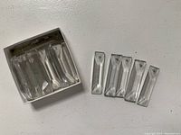 Seven tapered rectangular prism-shaped crystal chandelier drops, five loose and two inside a small white box. The crystals are clear and faceted, with small holes for hanging.
