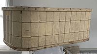 Side image of large antique laundry basket showing woven pattern, rounded corners, metal rivet reinforced top edge and solid build.