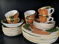Full set of vintage 1960s Vera Neumann Island Worcester dishes, showing stacked plates, bowls, and cups with hand painted orange pumpkin and green leaf design.