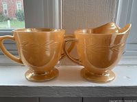 Photo showing the vintage Fire King cream pitcher and sugar bowl set in golden yellow/orange with visible leaf pattern detail and minor wear.