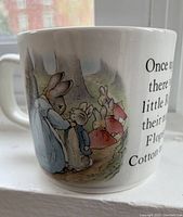 Side view of mug showing Peter Rabbit and sibling characters illustrated in watercolor style along with story text on white ceramic surface.