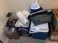 Lot showing disposable cups, plates, blue cooler boxes, insulated bag and other items stacked in corner.