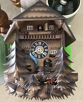 Front view of the wooden cuckoo clock showing carved pine tree details, clock face with Roman numerals, and figurine on balcony.