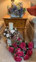Overall view of three faux flower arrangements including bright pink roses in front, white candle arrangement, and large multi-colored flower arrangement on table