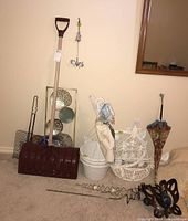 Photo showing large snow shovel with plastic red blade and wooden handle, assorted white metal and plastic hanging baskets with intricate designs, floral umbrella, and additional garden decorative items on a carpeted floor.