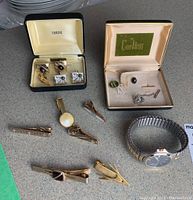 Photo showing multiple pieces of men's jewelry on a table including cuff links in cases, tie clips, and a wristwatch